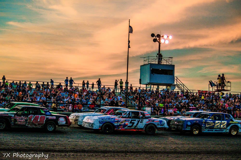 Stock cars lined up on a dirt track at sunset, with a crowded grandstand behind them.