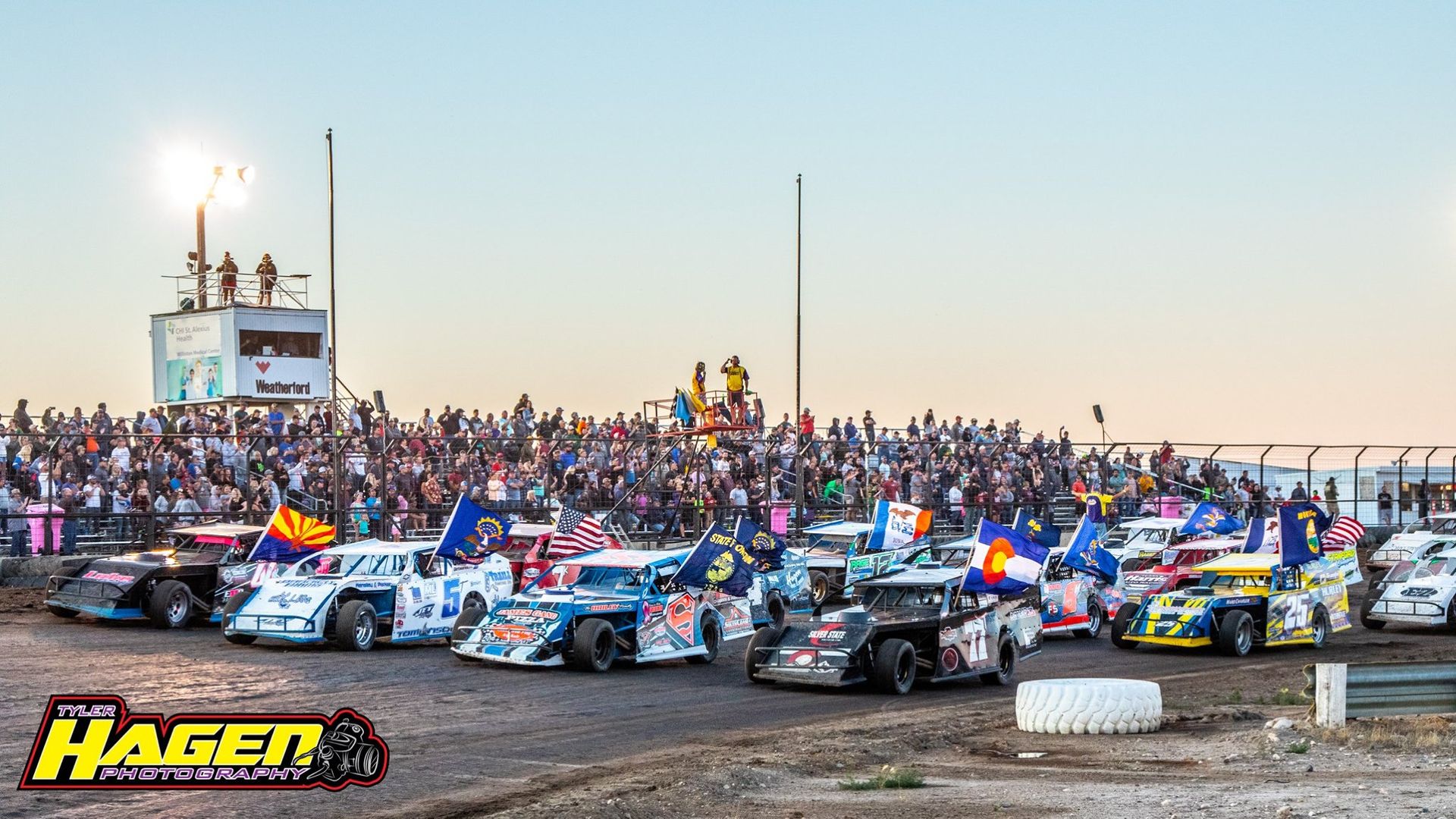 Dirt track race lineup with colorful stock cars before the start, packed grandstand in the background.