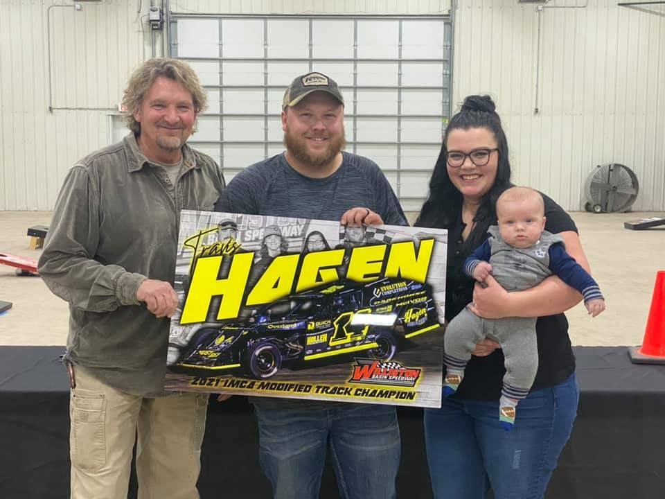 Four people holding a yellow-and-black racing sign inside a garage or workshop