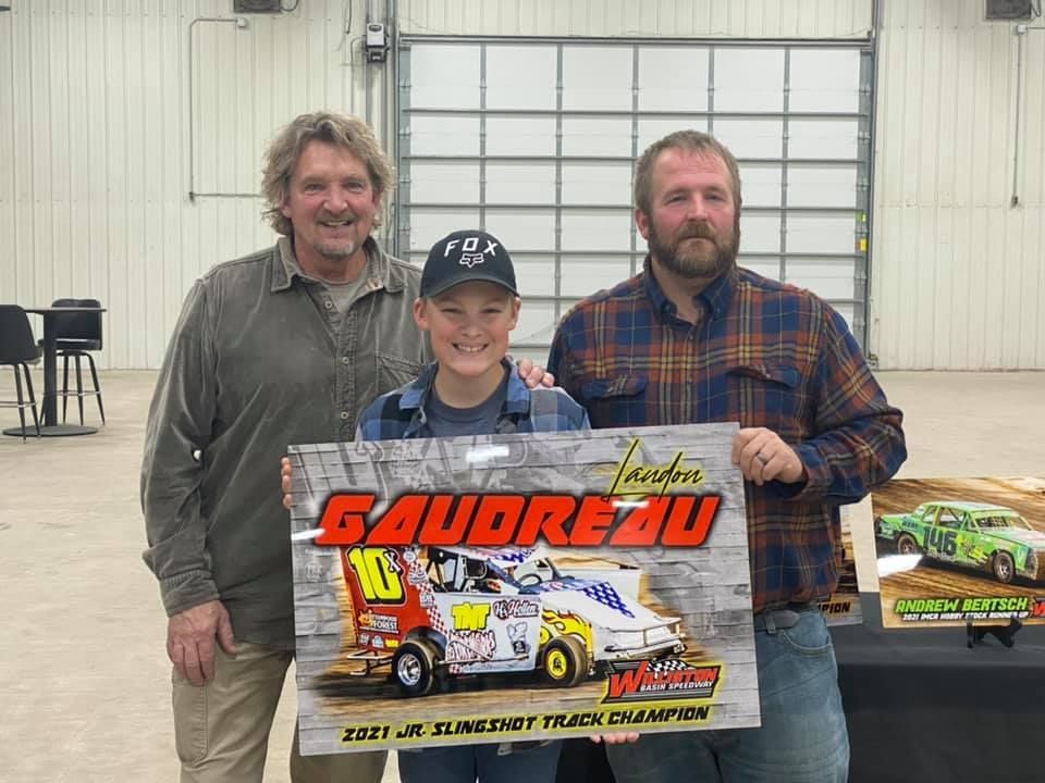 Three people hold a large Saubleau racing poster in a warehouse, smiling beside a race car photo.