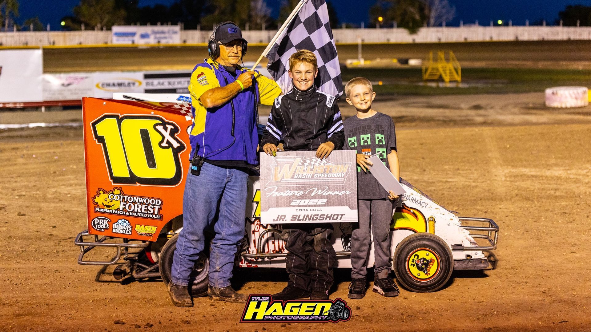 Three racers posing with trophy and checkered flag beside a dirt track race car at night
