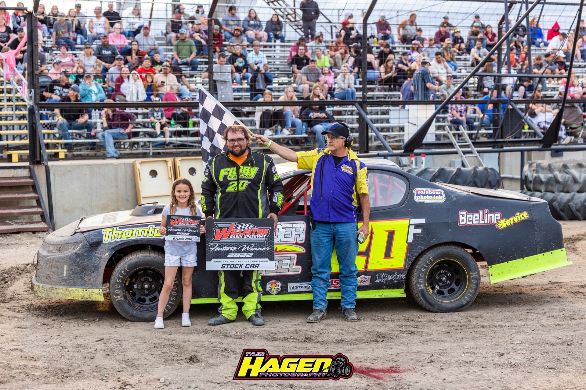 Three people posing beside a black race car on a dirt track with spectators in bleachers.