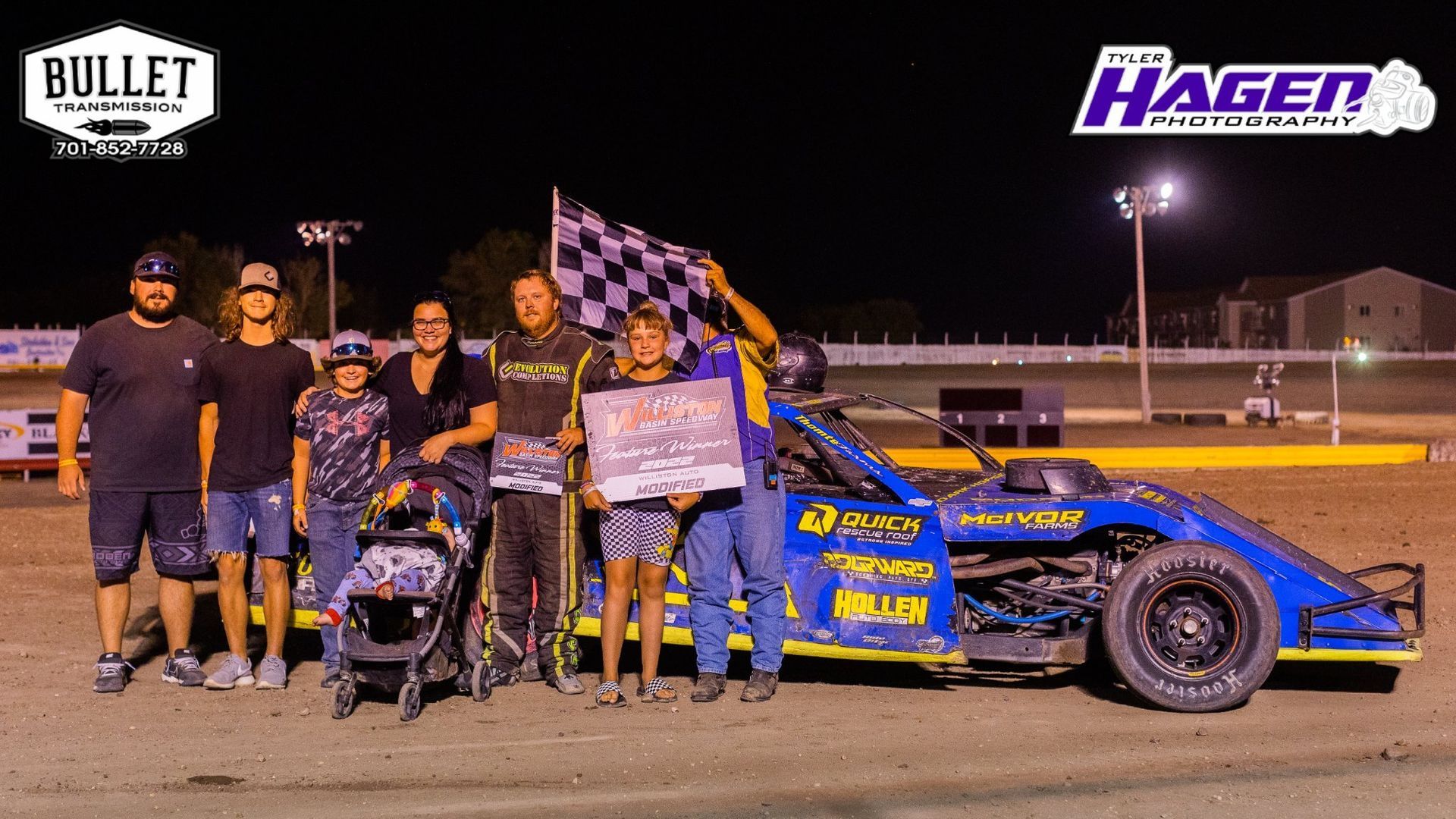 Race team posing beside a blue and yellow sprint car under lights, with trophy and checkered flag