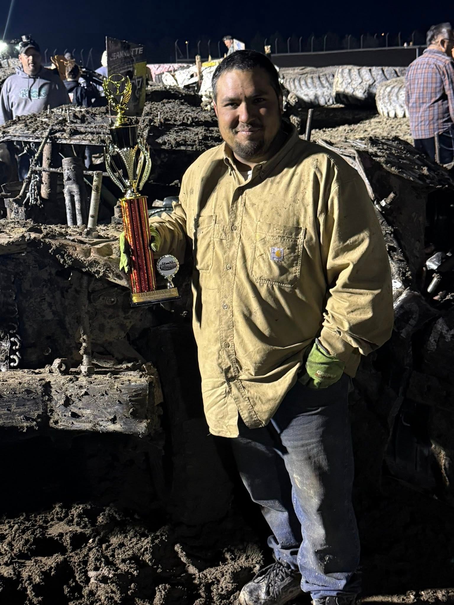 Man in a yellow shirt holding a drink at a muddy nighttime outdoor event with debris and people behind him