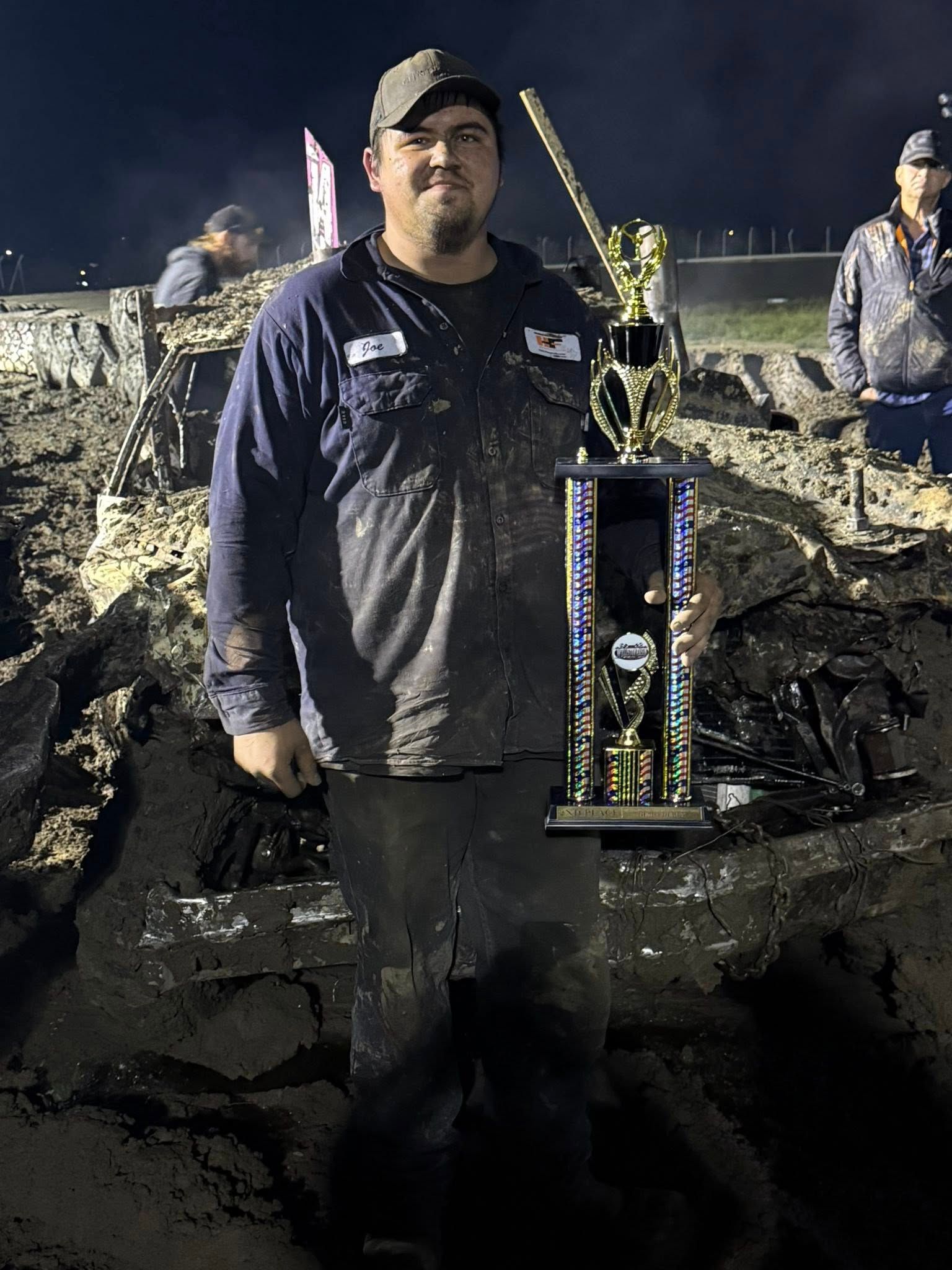 Person standing outdoors at night holding a trophy, with rubble and two people in the background.