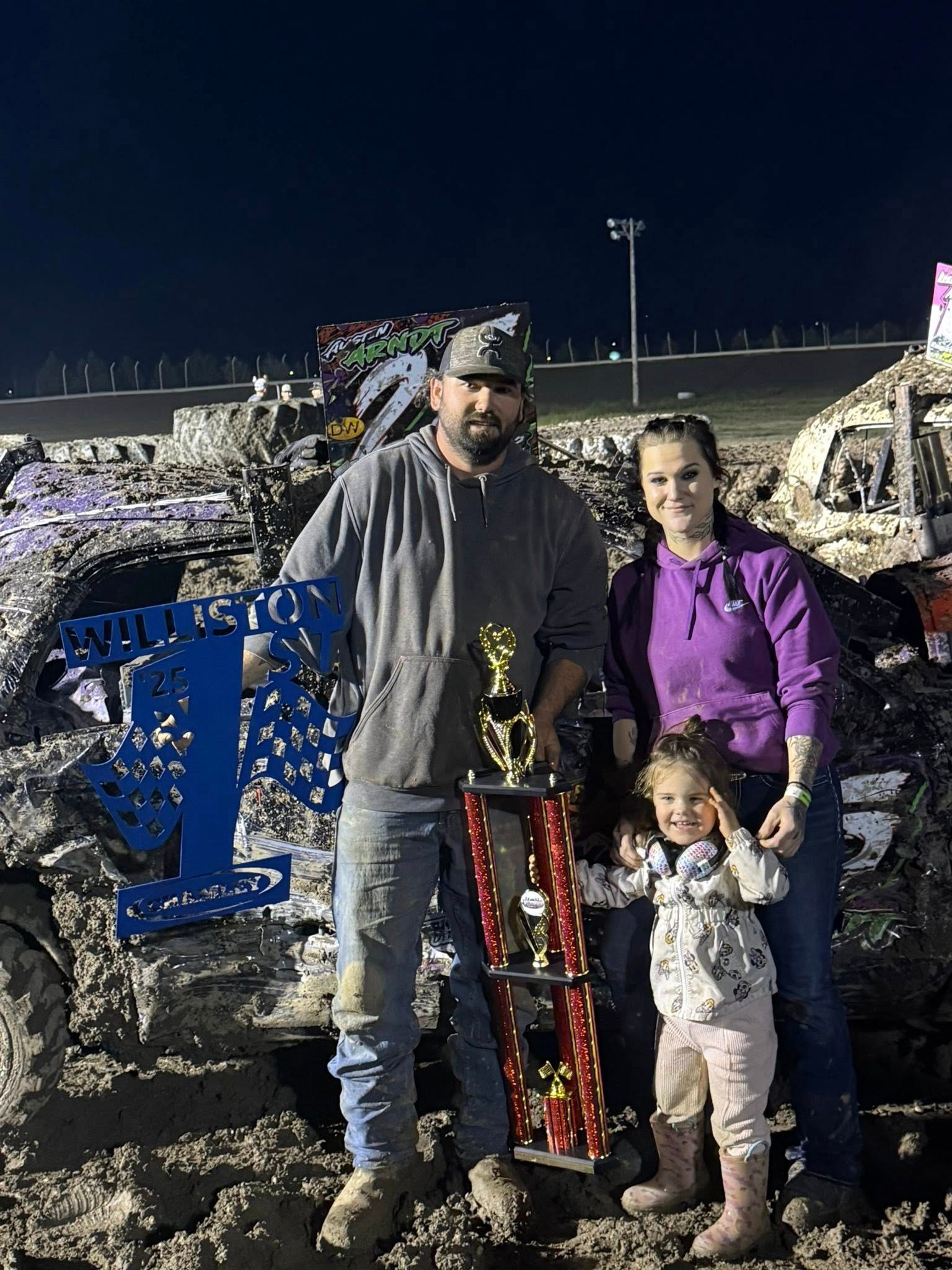 Man, woman, and child posing beside a red toy truck at a muddy nighttime outdoor event