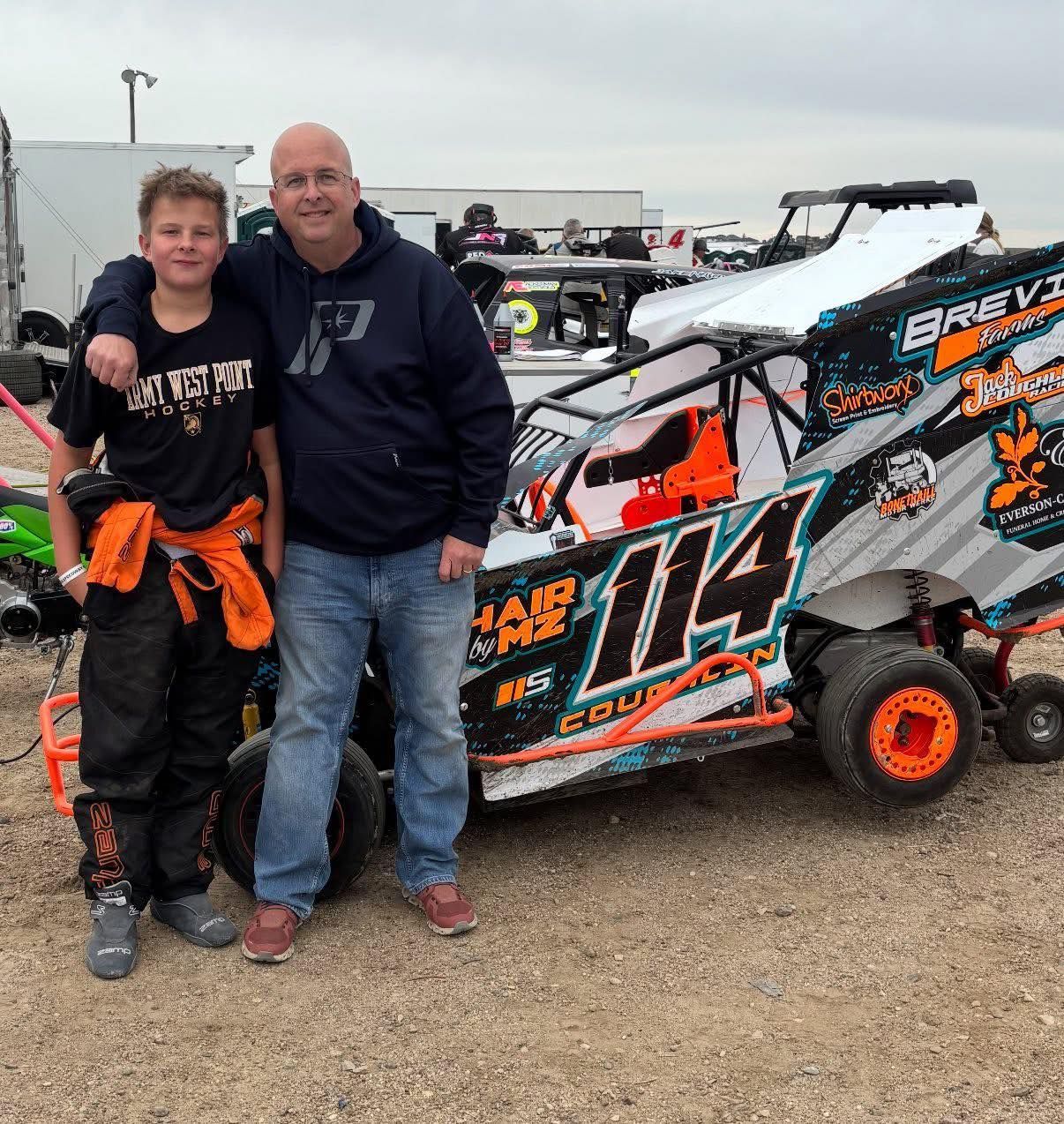 Two people standing beside a white and orange race car on a gravel lot
