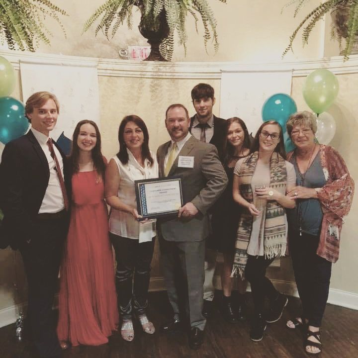 A group of nine people posing in a room with balloons and ferns, one person holding a framed award certificate.