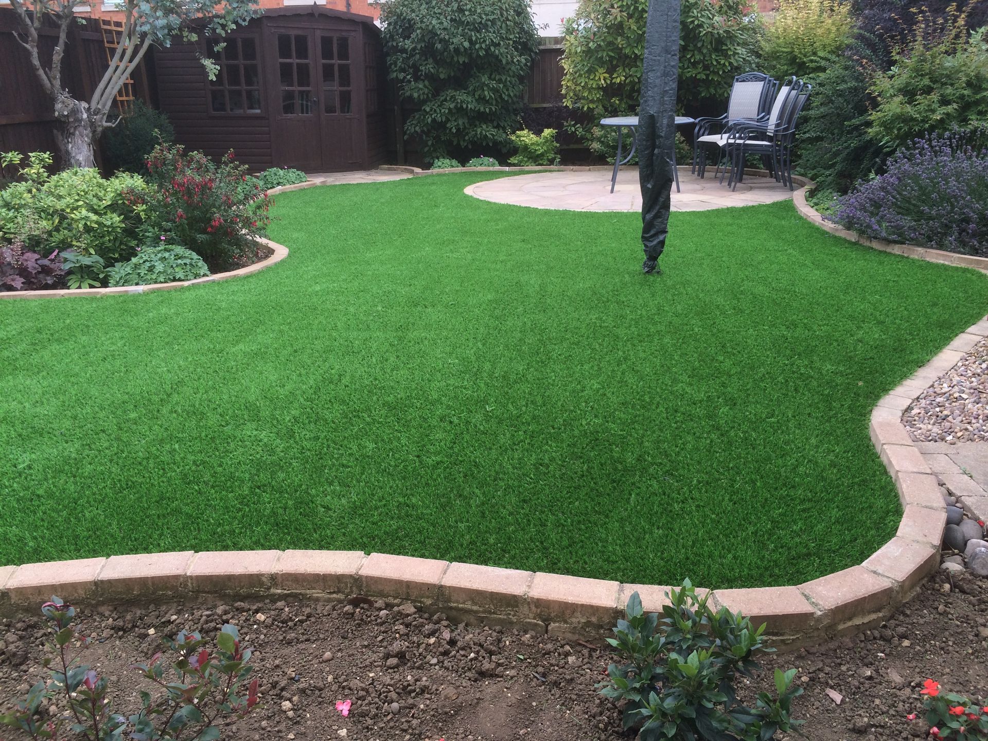 Green artificial lawn in a garden, edged with brick and bordered by plants.