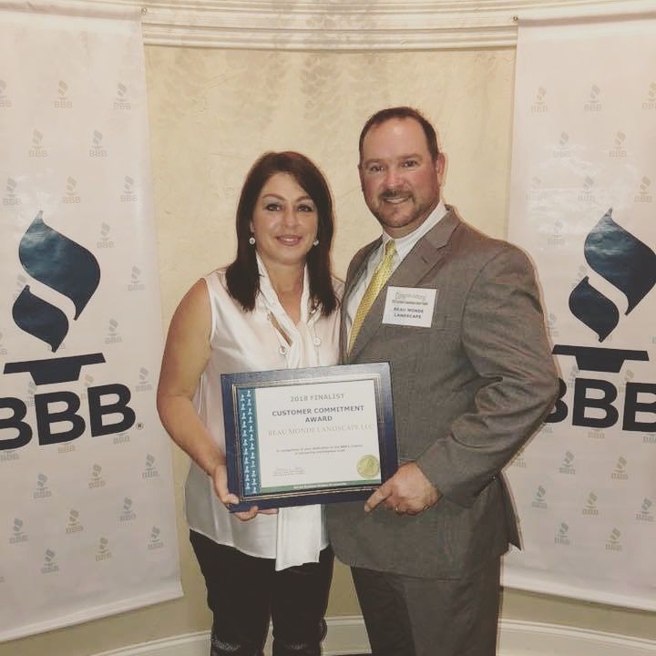 A woman and a man in business attire hold a Better Business Bureau award certificate in front of two BBB banners.