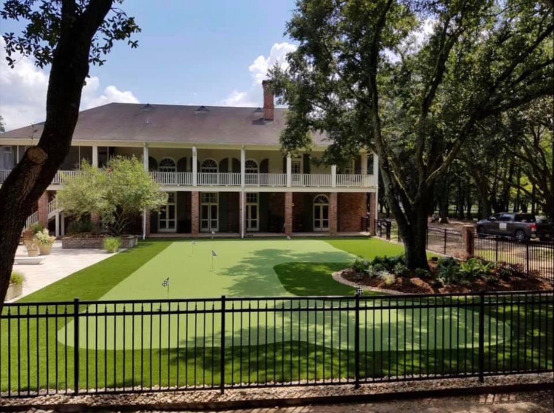 Large house with two-story porch, manicured lawn, putting green, black fence, and trees under a sunny sky.