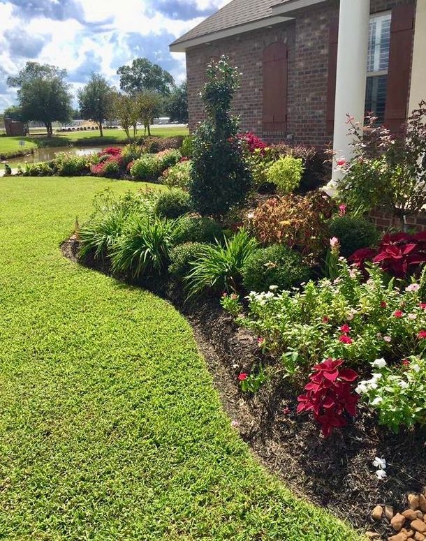 A manicured garden bed curves along a green lawn beside a brick house with white columns.