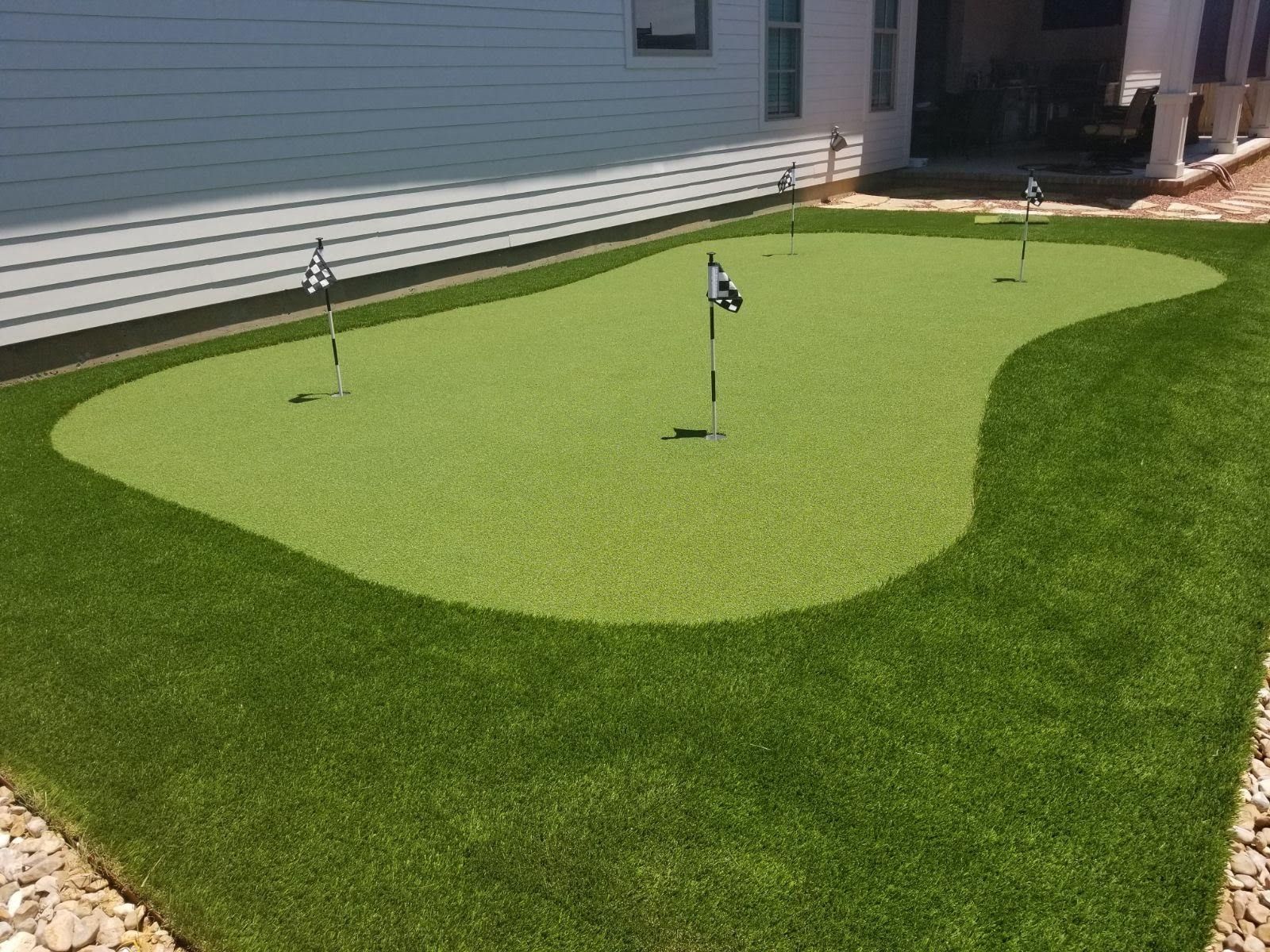 Artificial putting green with multiple holes, flags, and surrounding artificial turf next to a house.