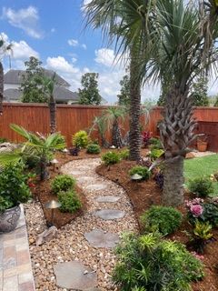 Curved stone path through a tropical garden with palm trees, shrubs, and a wooden fence under a blue sky