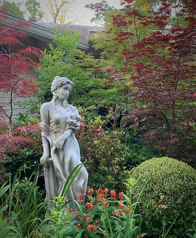 A stone statue of a figure stands in a lush garden filled with green and red maple trees and small red flowers.