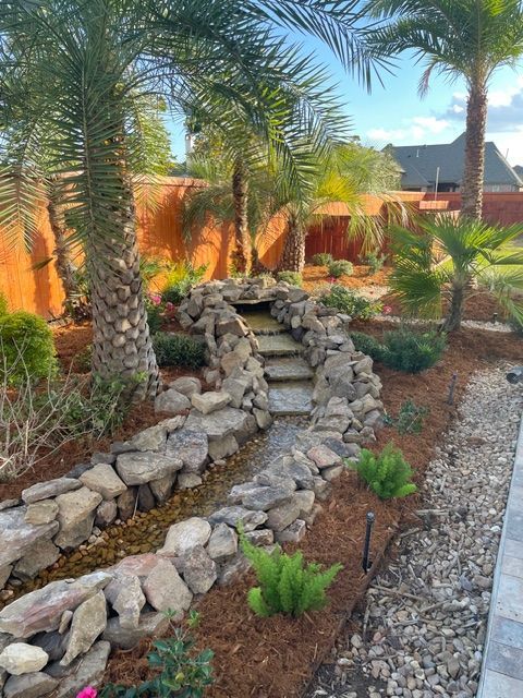 A stone water feature winds through a landscaped backyard with palm trees, mulch, and a gravel border.