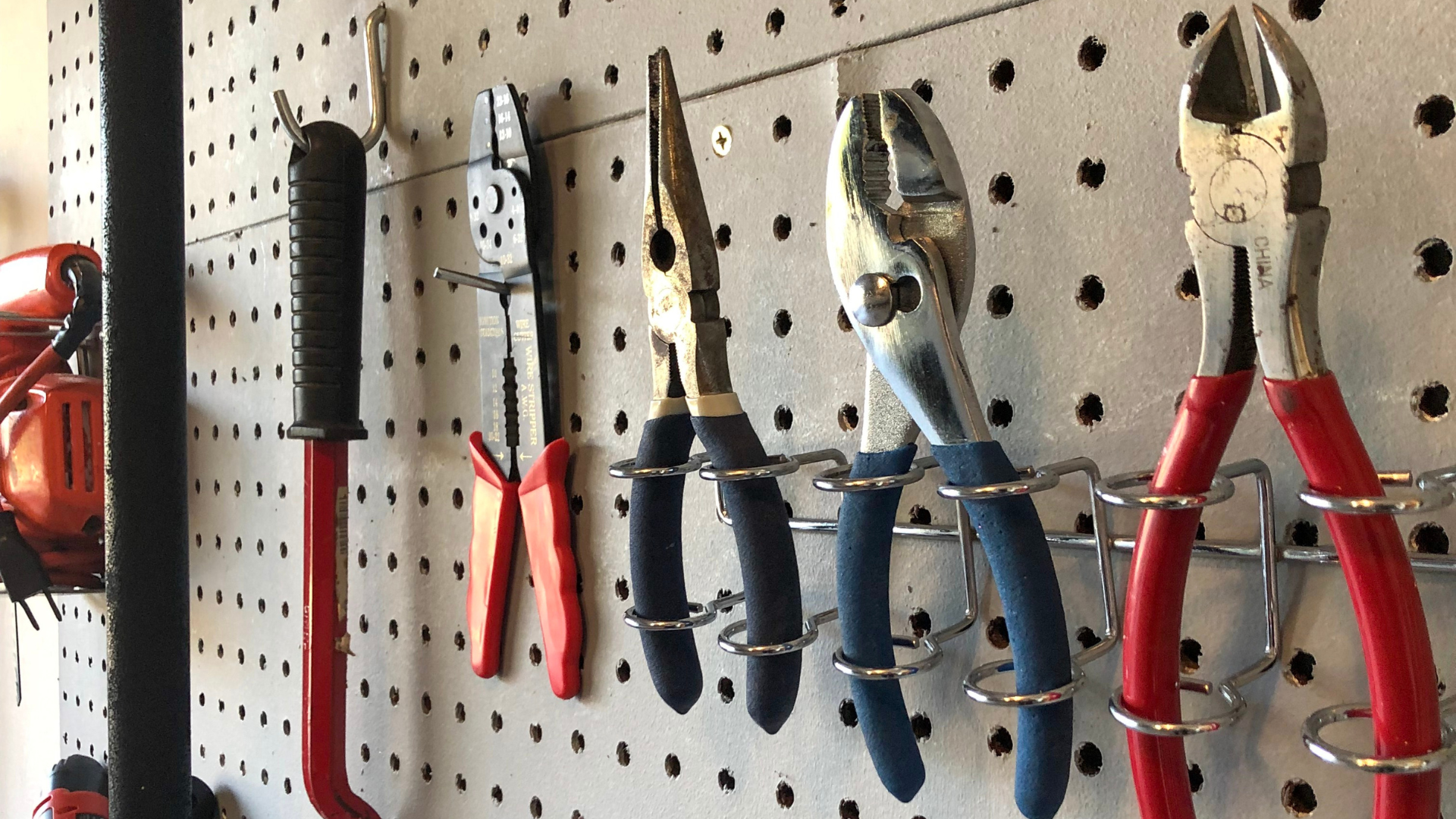 Tools hanging on a pegboard: pliers with red, blue, and black handles, and a red bar.