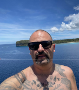 A shirtless man wearing sunglasses takes a selfie in front of the ocean