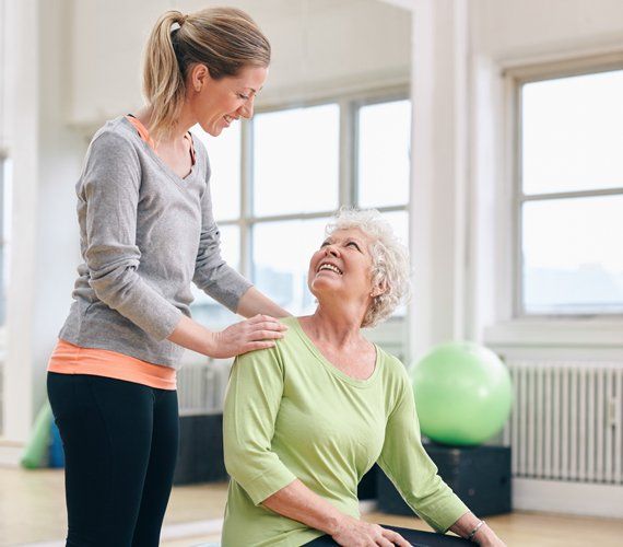 Woman assisting another woman stretching indoors, smiling.