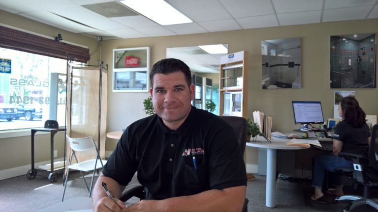 Man in black shirt smiles in an office, desk, window, and another person at a computer are visible.