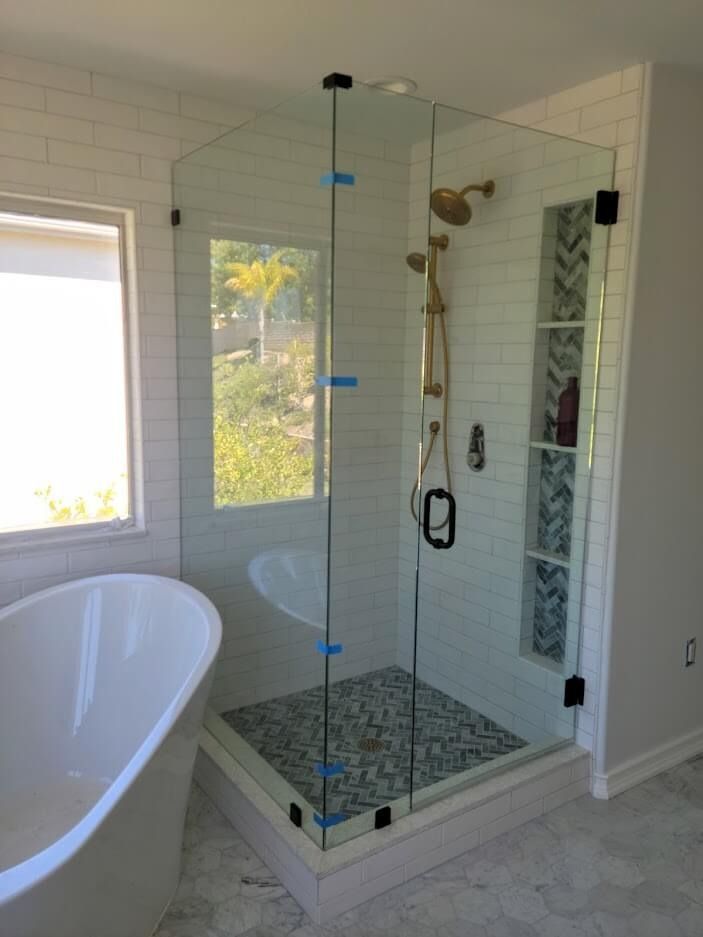 A glass shower enclosure with blue-tiled floor and gold fixtures, next to a white bathtub.