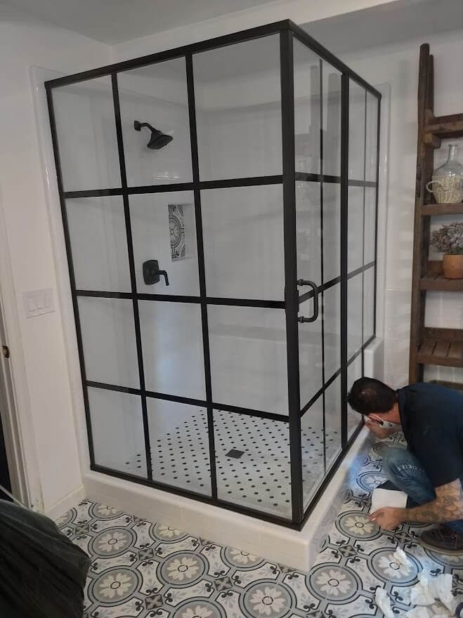 A person installing tile near a modern shower enclosure with a black grid frame.
