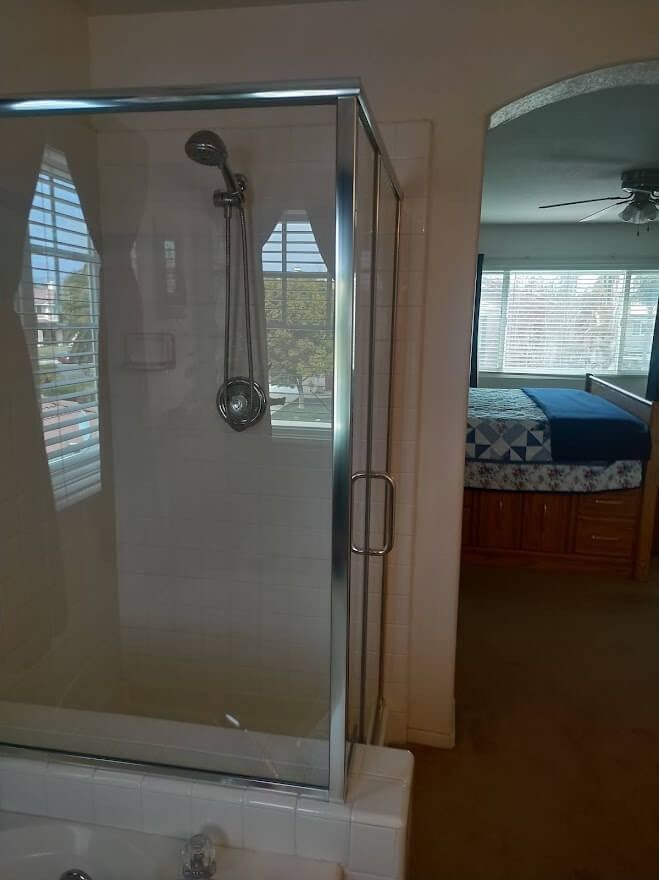 Shower with glass door, next to a bedroom with bed, white walls, and bright sunlight.