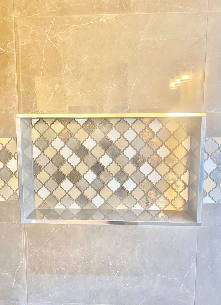 Shower niche with mosaic tile design, framed by silver trim, in a tiled wall with neutral colors.