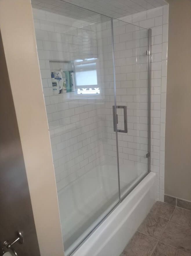 Glass shower door on a white tiled wall, over a bathtub with a beige wall beside it.