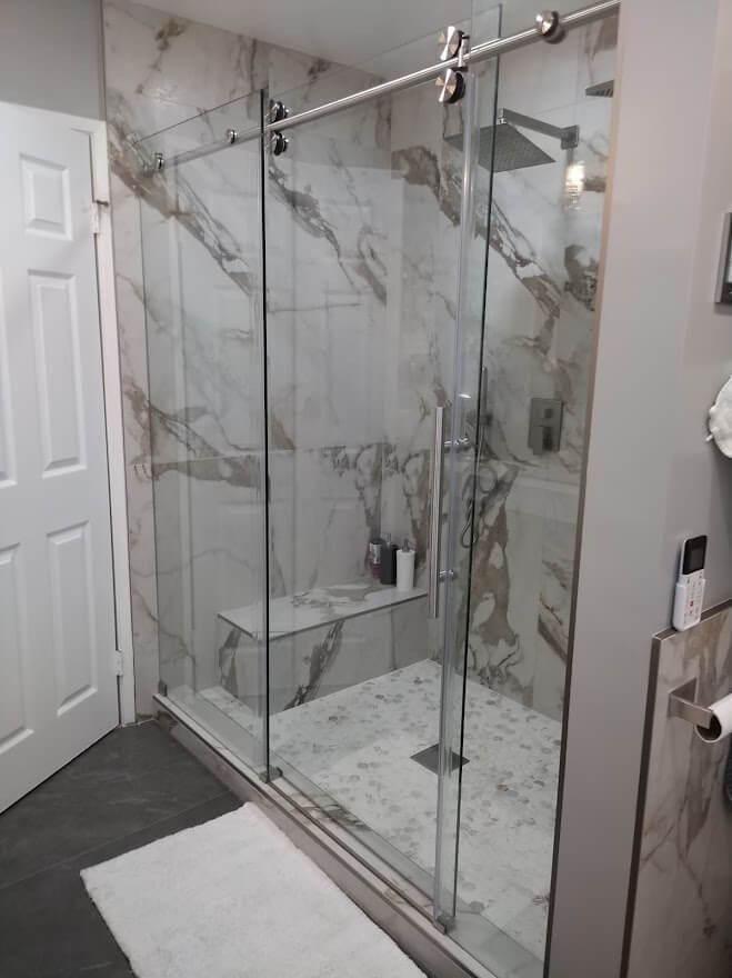 Modern bathroom with glass shower doors and marble-like tile walls. White door on left, gray walls, white rug.