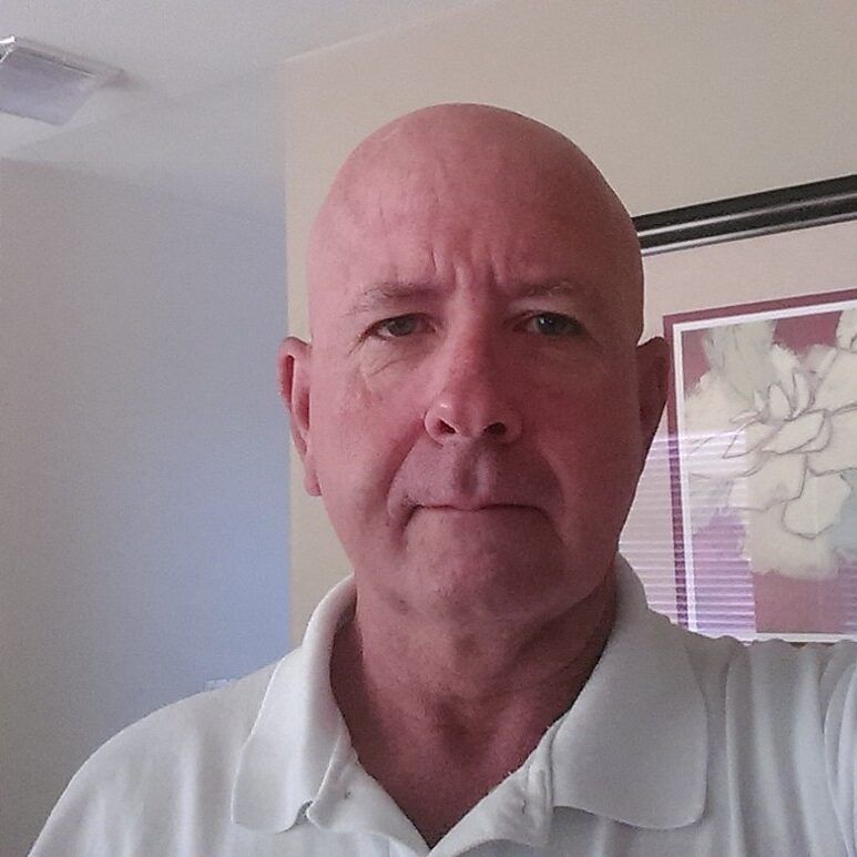 Bald man in a white collared shirt, looking at the camera indoors, neutral expression.