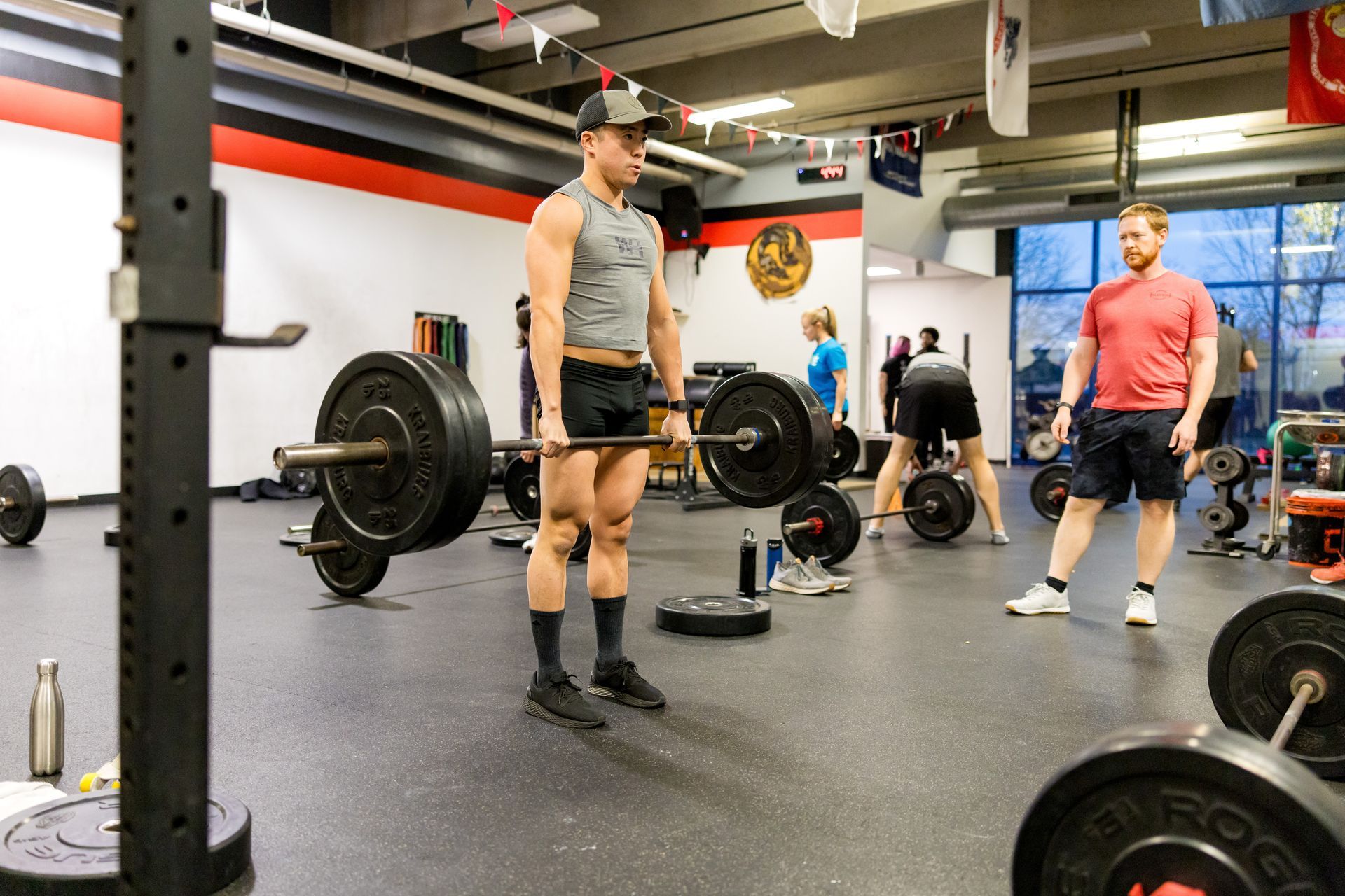 Man lifting barbell in a gym; others exercising in background.