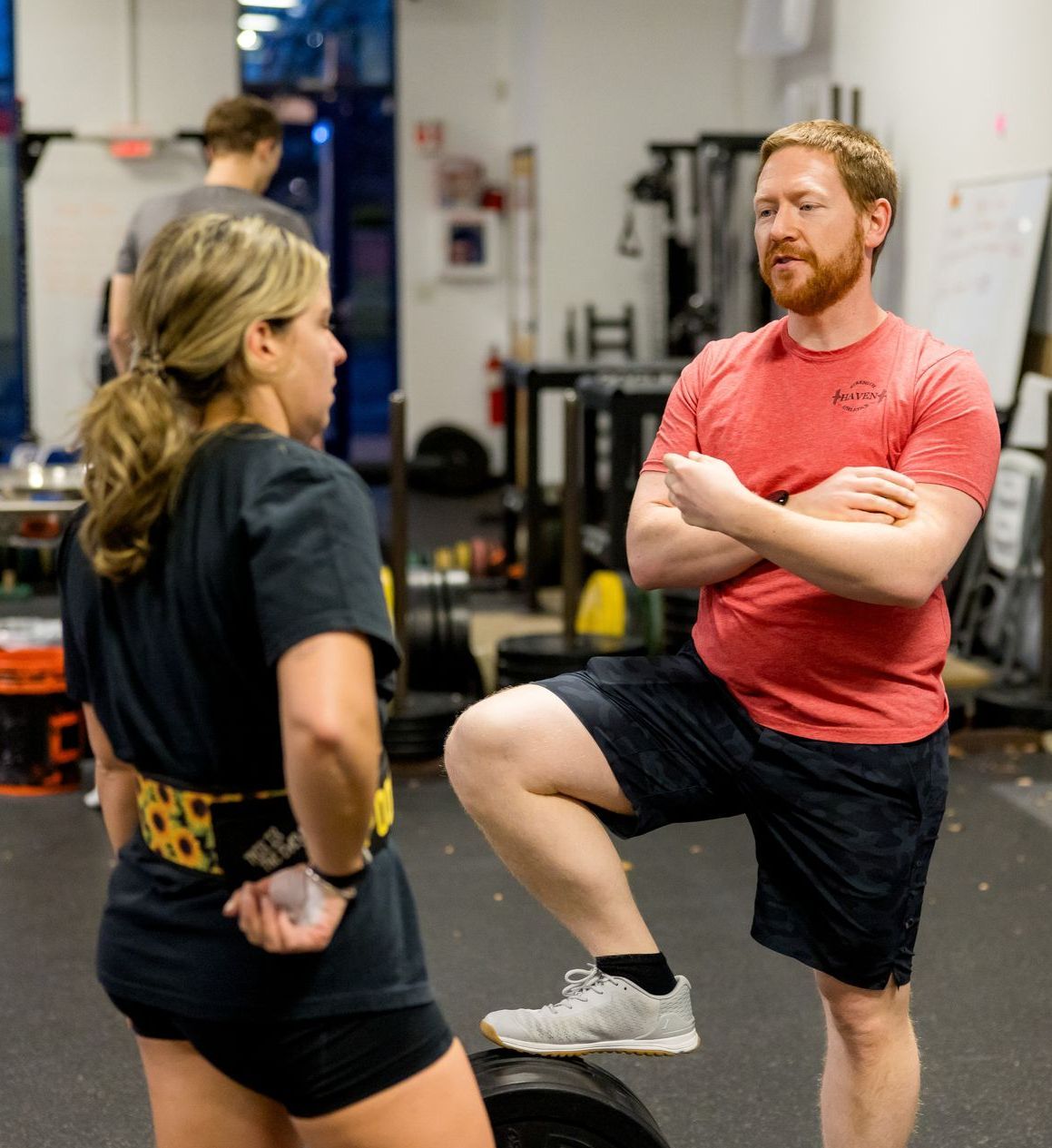 Woman and man in gym discuss strategy. Man rests leg on weights, arms crossed.