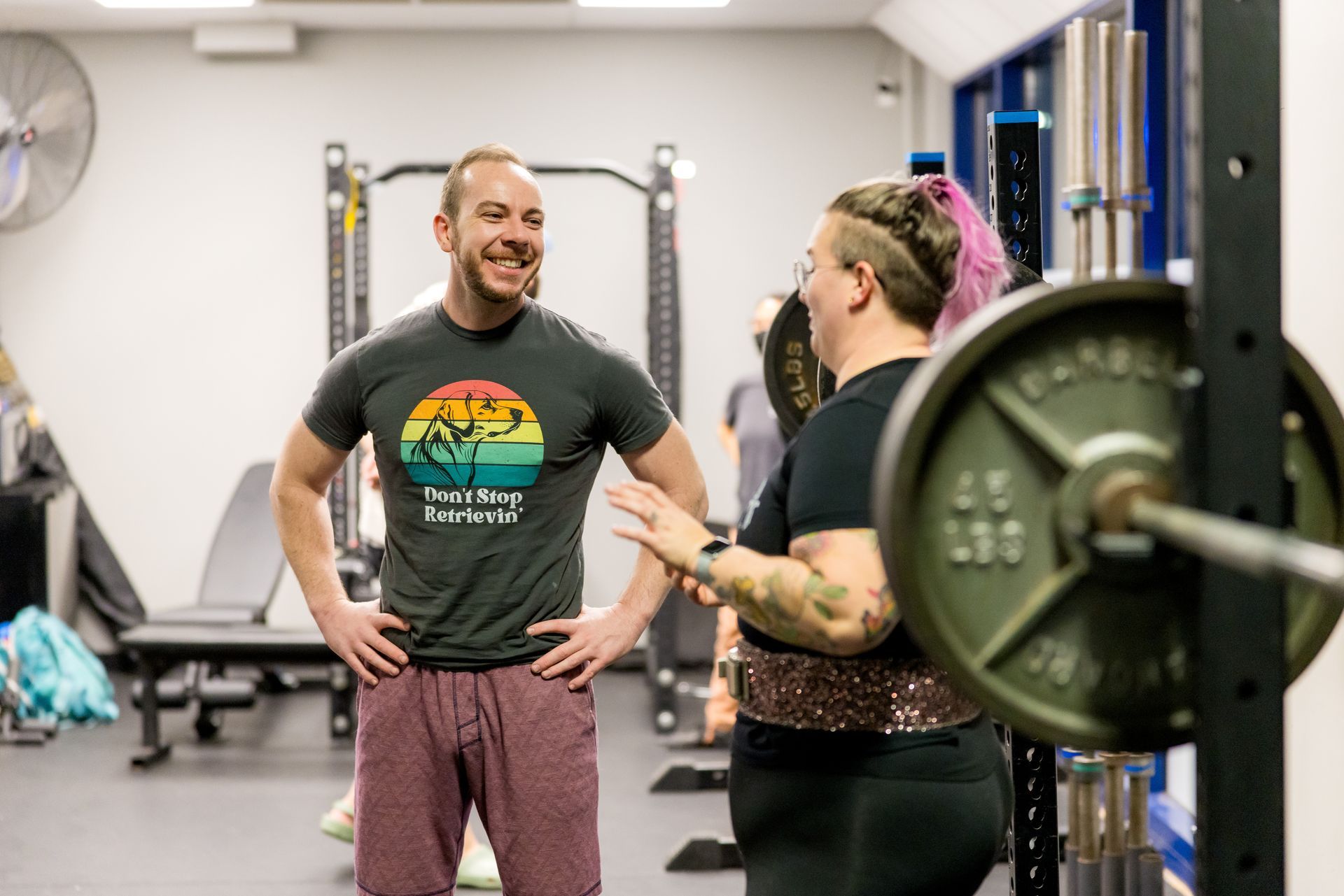 Man and woman at a gym, smiling. Woman has arm tattoos and weight on the bar.
