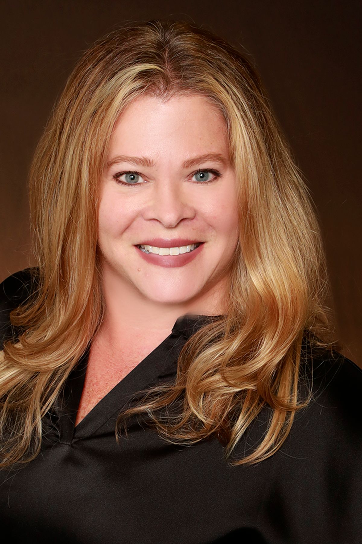 Woman with wavy blonde hair, smiling, wearing a black shirt, against a brown background.
