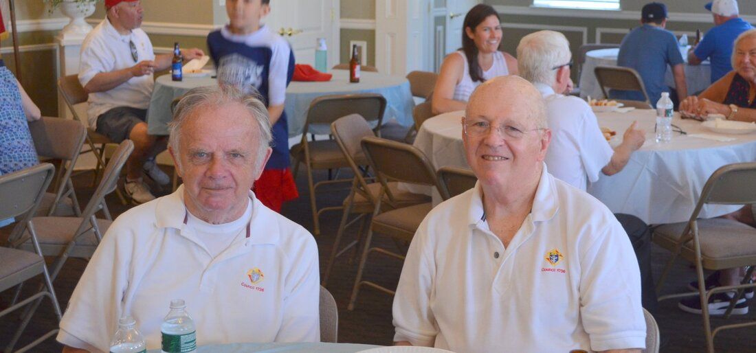 Two older men are sitting at a table in a room with other people.