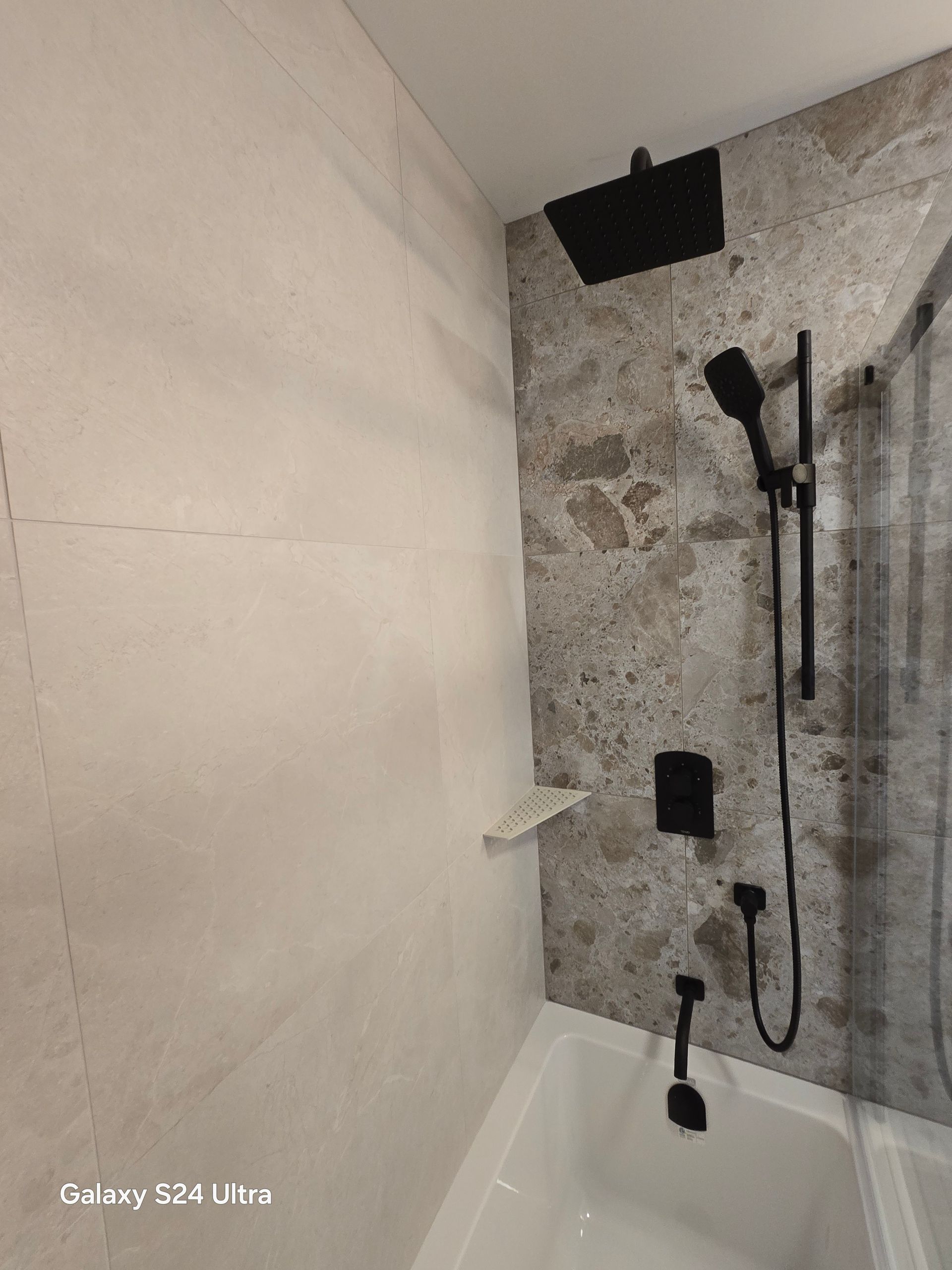 Douche avec robinetterie noire et carrelage imitation pierre. Comprend une baignoire, une pomme de douche effet pluie et une douchette.