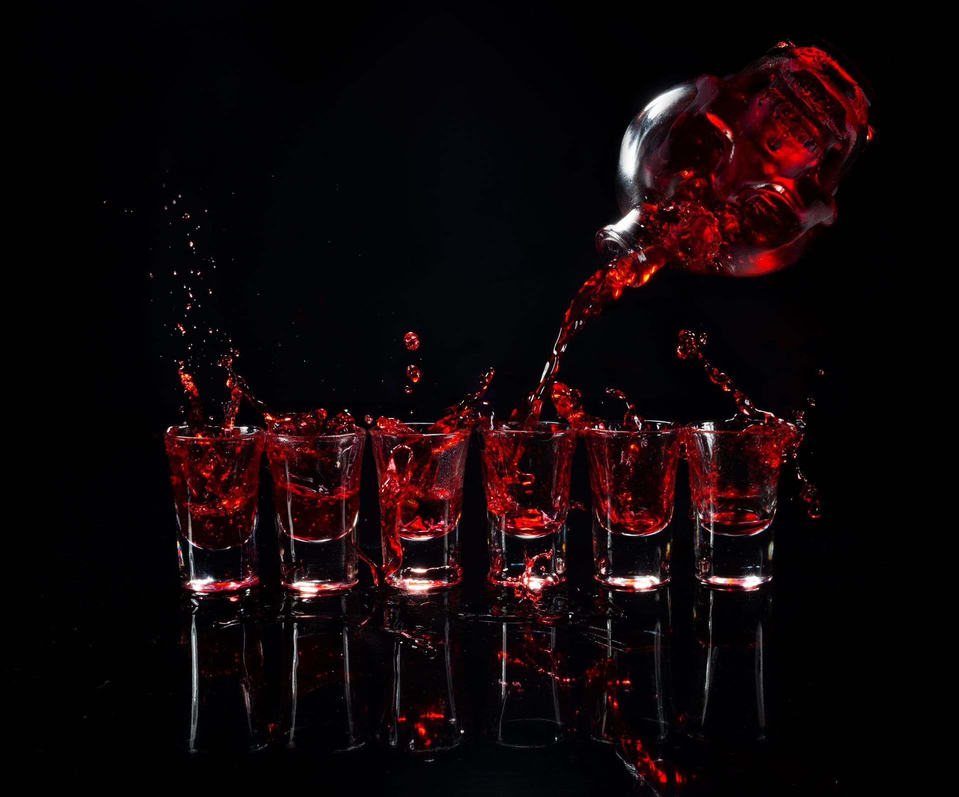 Eine blutrote Flüssigkeit wird in sechs Pinnchen gegossen. Der Hintergrund ist schwarz.