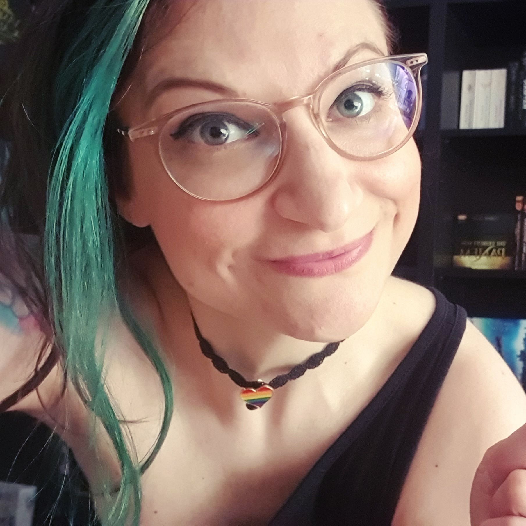 Lächelnde Frau mit geschminkten Augen, Brille und grün gefärbtem Haar. Um den hals trägt sie eine Kette mit Regenbogen-Herz