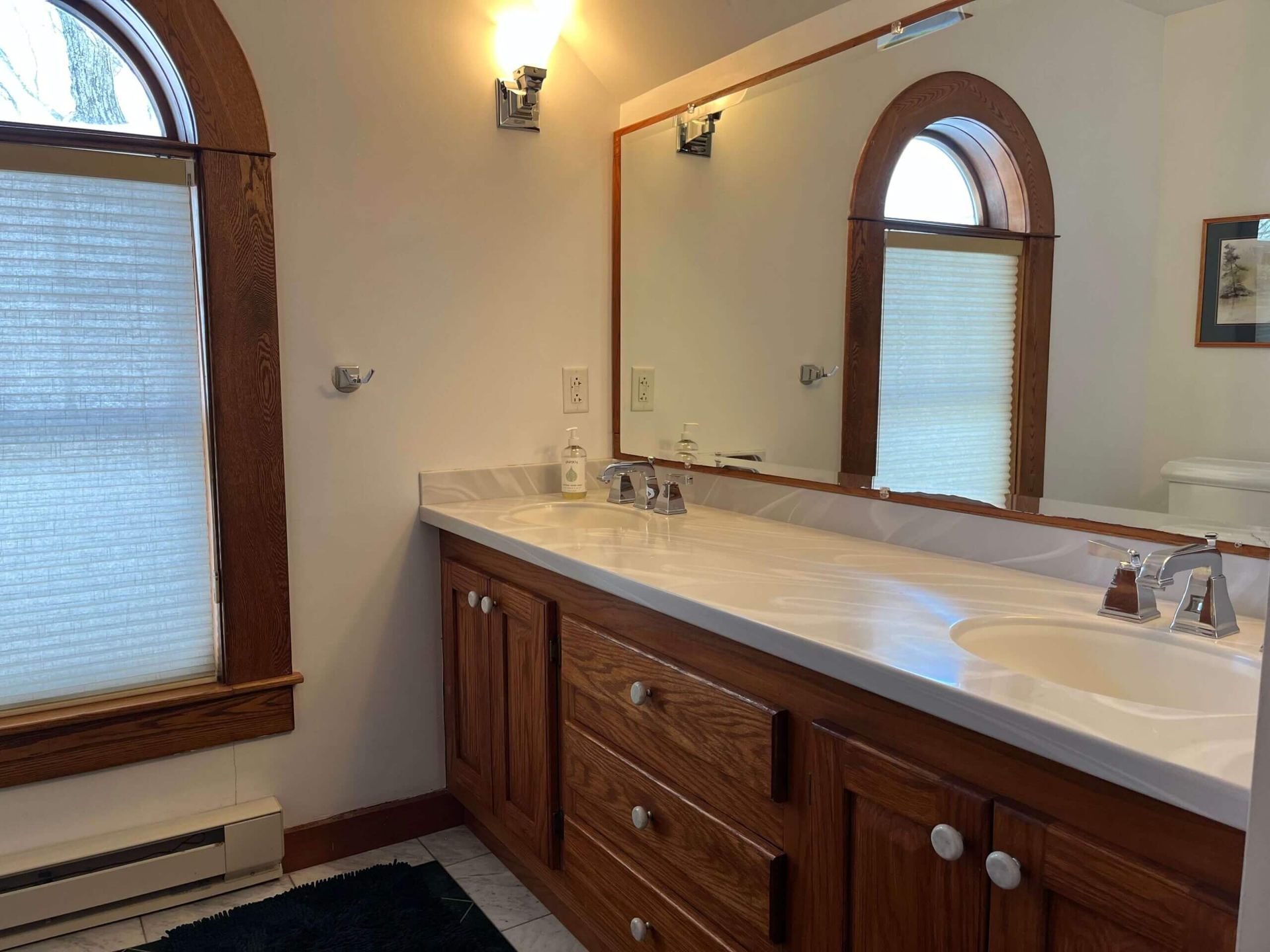 Bathroom with a long vanity, wooden cabinets, arched window, and large mirror.