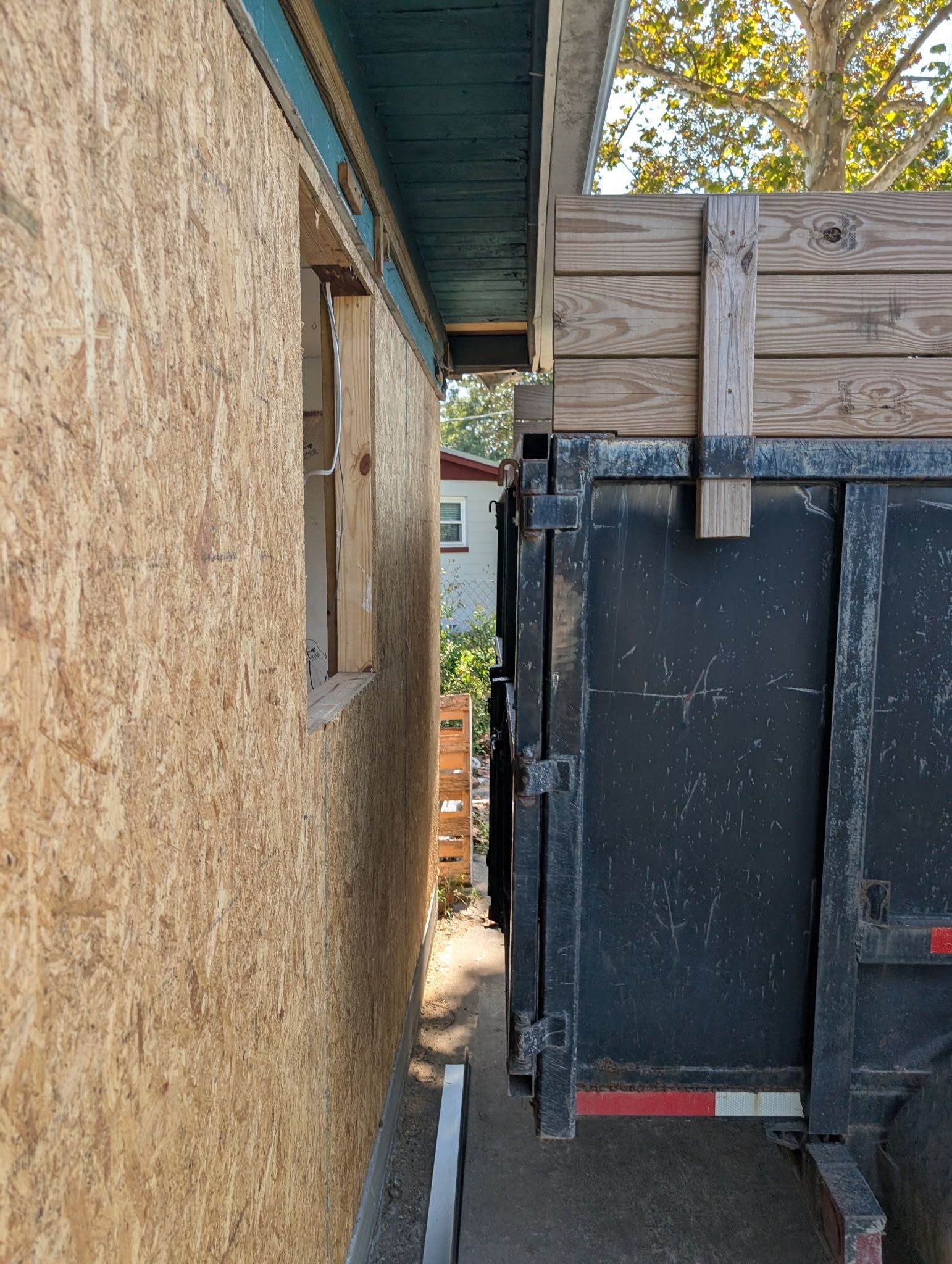 A green dumpster is sitting next to a pile of wood in front of a house.