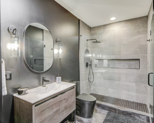 Modern bathroom with a wooden vanity, oval mirror, wall sconces, and a glass-enclosed shower with gray tiled walls.