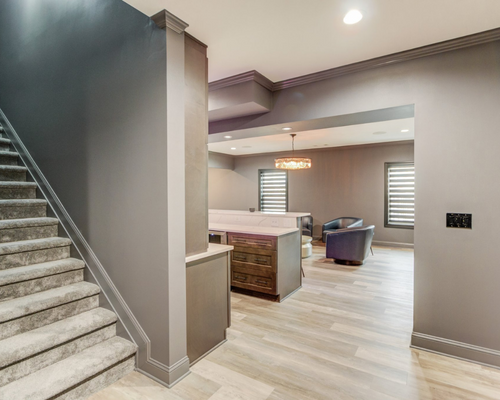 A staircase with grey carpet leads to an open-plan room with grey walls, wooden flooring, a kitchen island, and chairs.