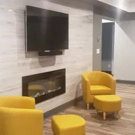 A modern living area with a wall-mounted TV above a fireplace, featuring two yellow armchairs and matching ottomans.