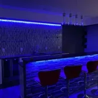 A dimly lit home bar with a blue LED-lit counter and brick textured wall, featuring three red stools and pendant lighting.