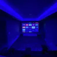 A darkened home theater room illuminated by blue cove lighting, featuring a large screen displaying media app icons.