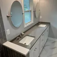 A modern bathroom vanity with a long gray countertop, two white rectangular sinks, oval pivoting mirrors, and white cabinets.