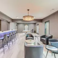 A modern, minimalist lounge featuring a long bar with stools, a central chandelier, and grey seating areas.