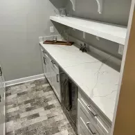 A built-in bar area featuring white marble countertops, gray cabinetry, tiled floor, and floating shelves.