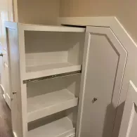 A white pull-out pantry unit with three empty shelves, partially extended from an alcove under a staircase.